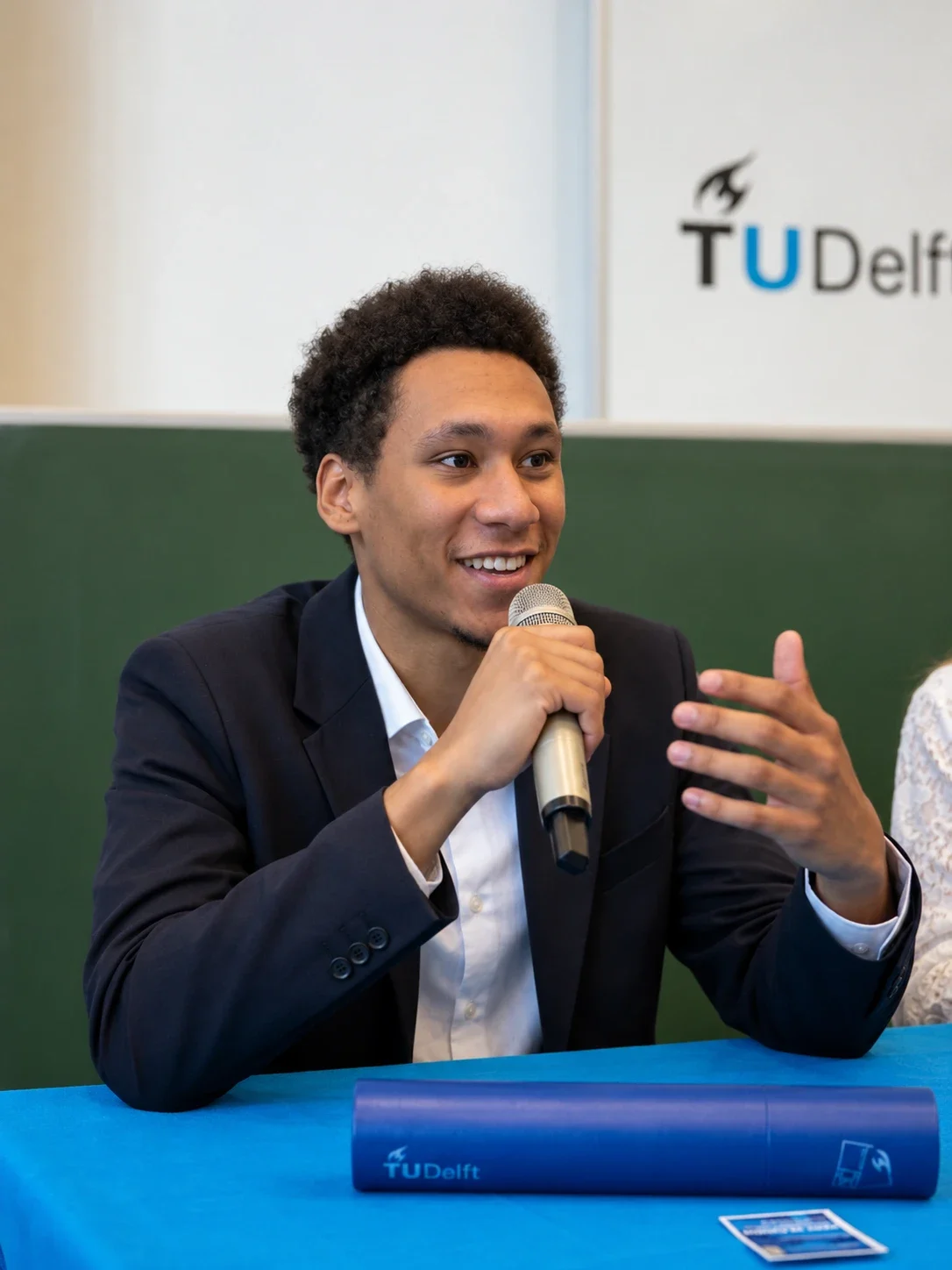 Wilbert Andrea aan het woord met een microfoon, in een donker colbert, bij de TU Delft